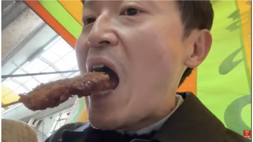 串に刺さった揚げ物を口元に運ぶ男性。