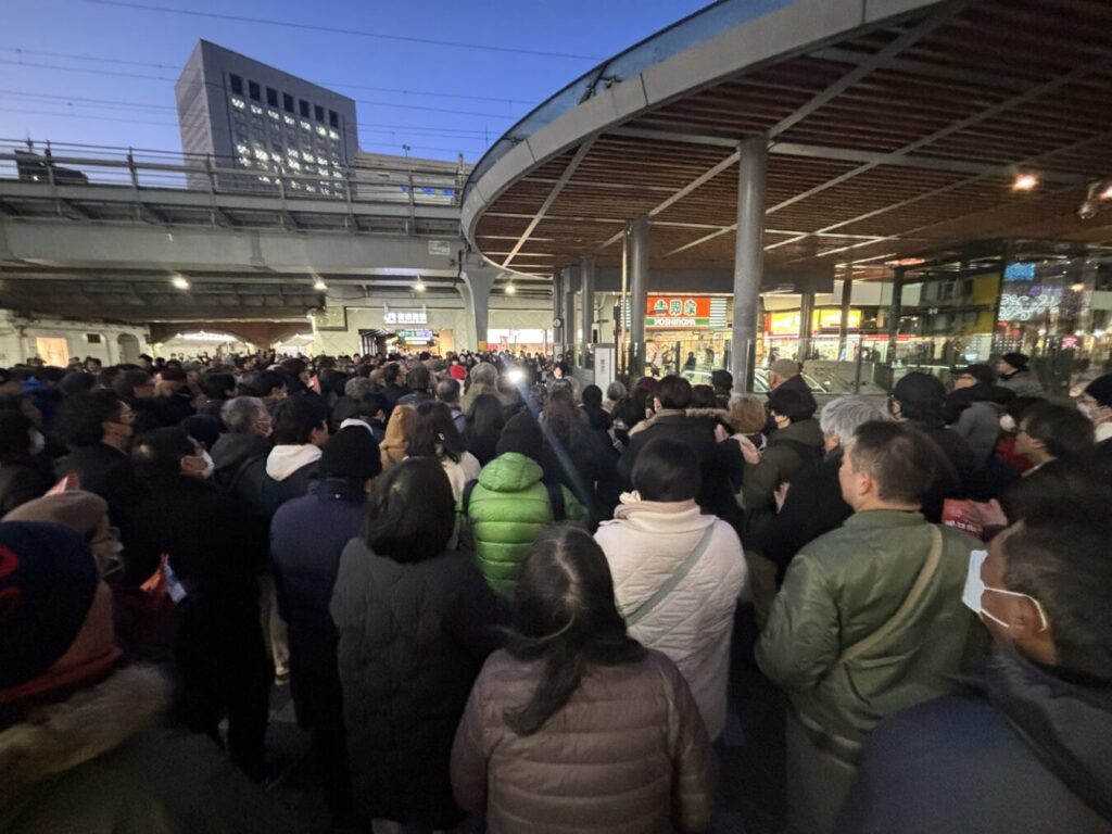 夕暮れ時の駅前広場に多くの人々が集まり、高架下や店舗周辺が混雑している様子。