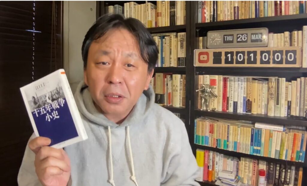 本棚を背景に、『十五年戦争小史』という本を手に持って話す菅野完。