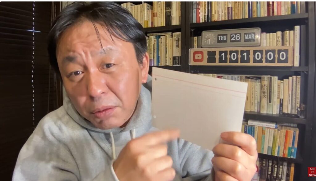 本棚を背に、白い無地の紙を手に持ち指差している菅野完。