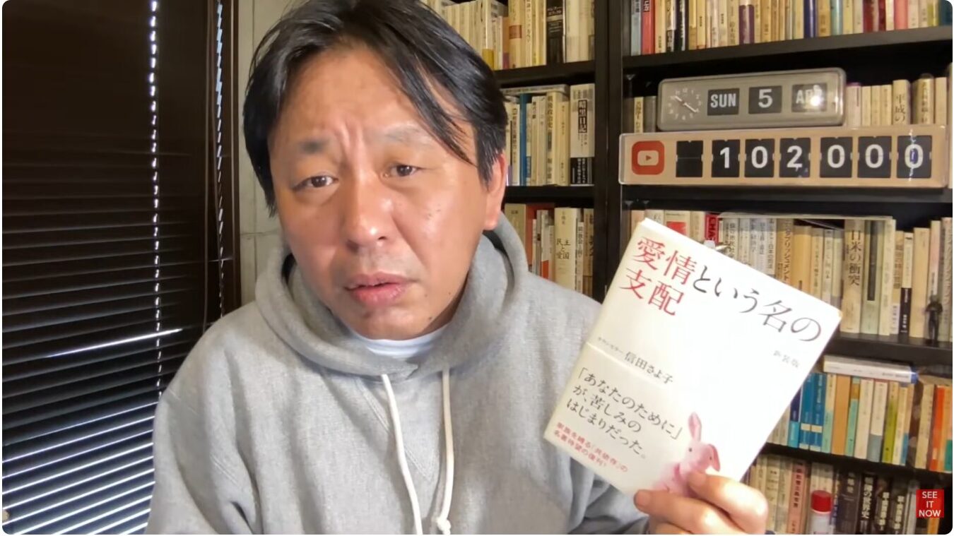本棚を背景にグレーのパーカーを着た菅野完が、信田さよ子著『愛情という名の支配』という書籍を手に持ってカメラを見ている様子。