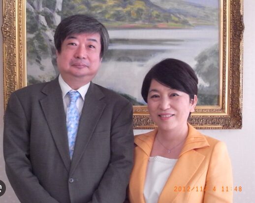 絵画を背景に並んで立つ、スーツとネクタイ姿の海渡裕一と、黄色のジャケットを着用した福島みずほのツーショット。