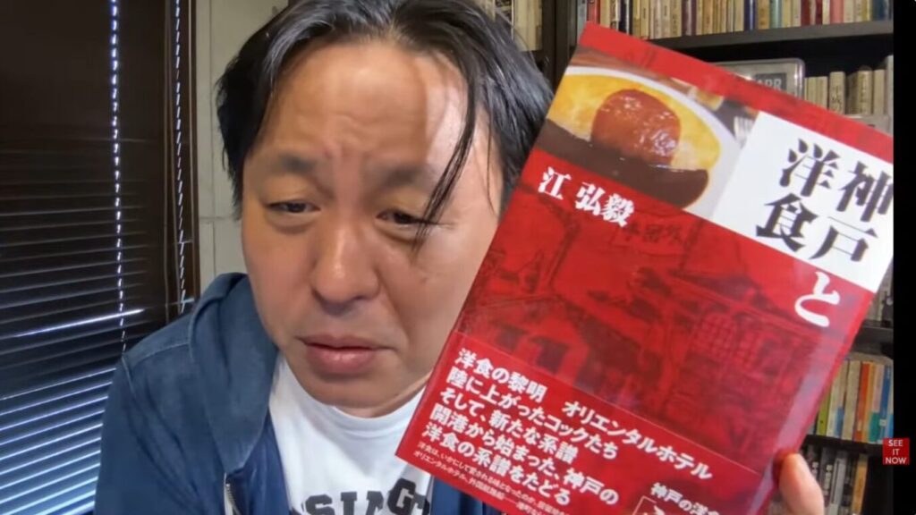 江弘毅著『神戸と洋食』を手に持ち、解説する菅野完。表紙にはオムライスのような料理や洋食の歴史に関する紹介文が記されている。