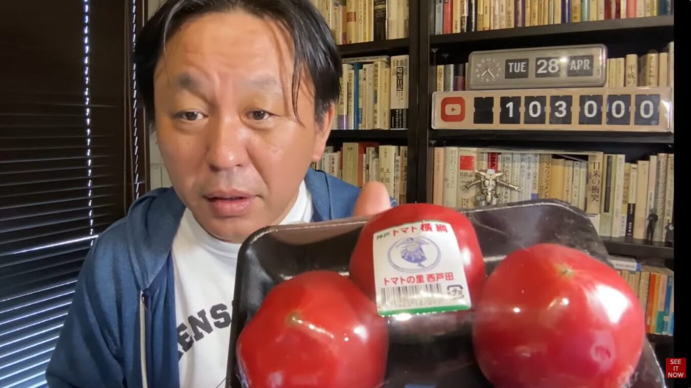 書棚を背景に、手前にトマト横綱と記されたラベルが貼られたパック入りの真っ赤なトマトを掲げる菅野完。