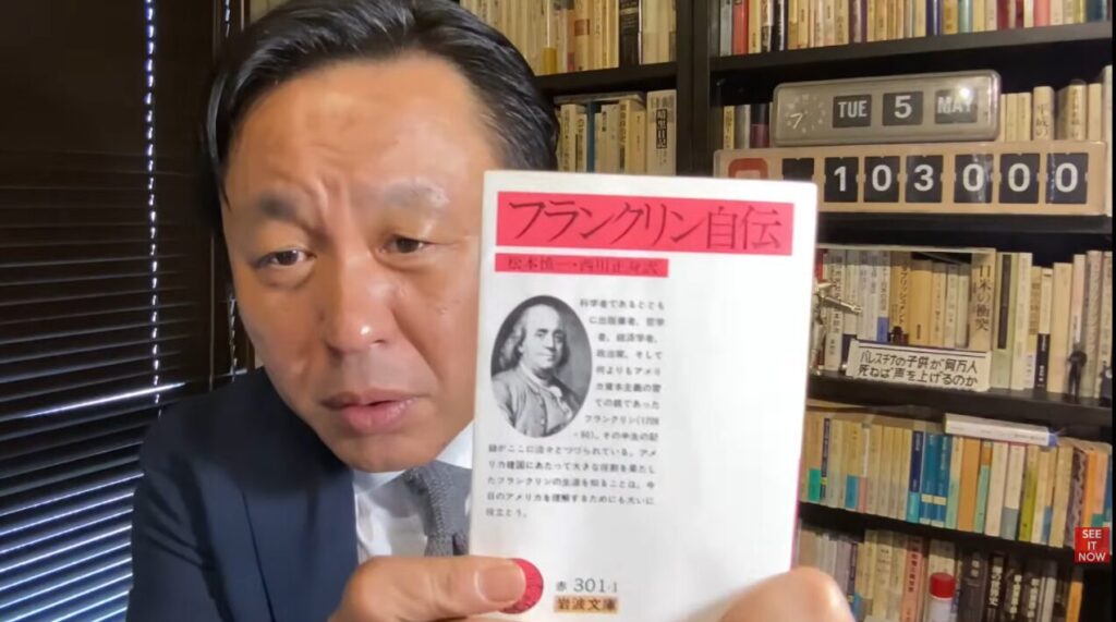 書棚を背景に、スーツ姿の菅野完が岩波文庫『フランクリン自伝』を手に持ち、表紙の解説文を提示している様子。