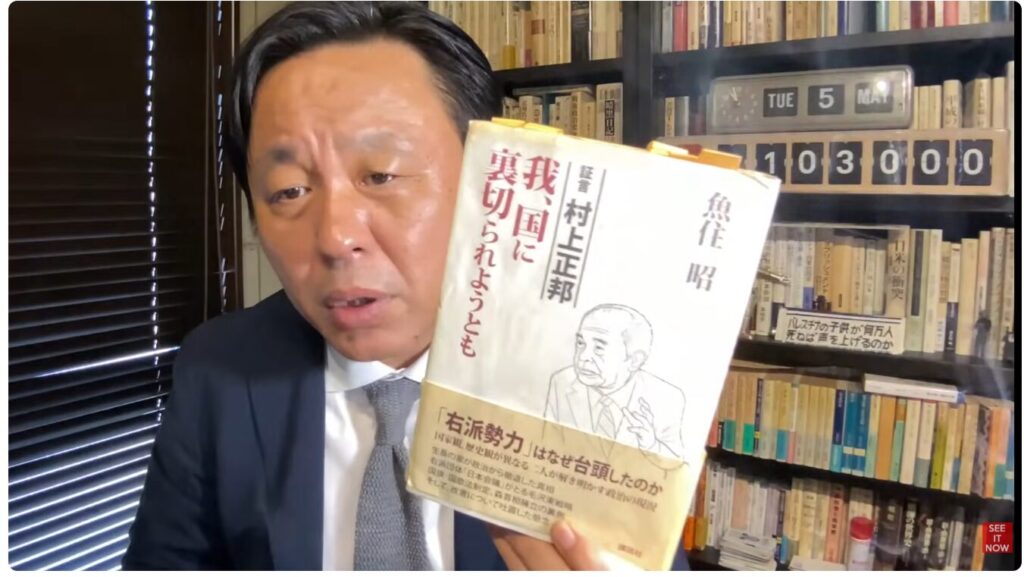 スーツ姿の菅野完が、村上正邦の著書『我、国に裏切られようとも』を手に持ち、正面に向けて解説している。