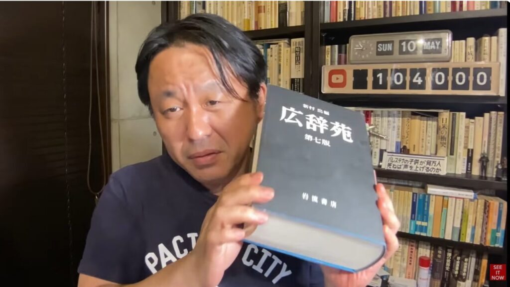 本棚を背景に、手元で『広辞苑 第七版』を掲げて視聴者に語りかける菅野完の様子。