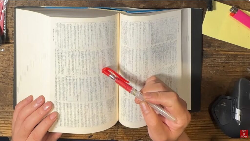 机の上で開いた辞書のページを赤いペンで指し示し、特定の語句を確認している様子。