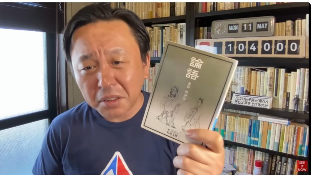 本棚を背にした菅野完が、手元で『論語』(金谷治訳注、岩波文庫)の表紙をカメラに向けて示している様子。