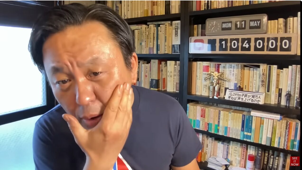 多くの書籍が並ぶ書棚の前で、顔に手を添えて語りかける菅野完。背景の棚には104000と示されたカウンターとパレスチナ問題に関する書籍が置かれている。