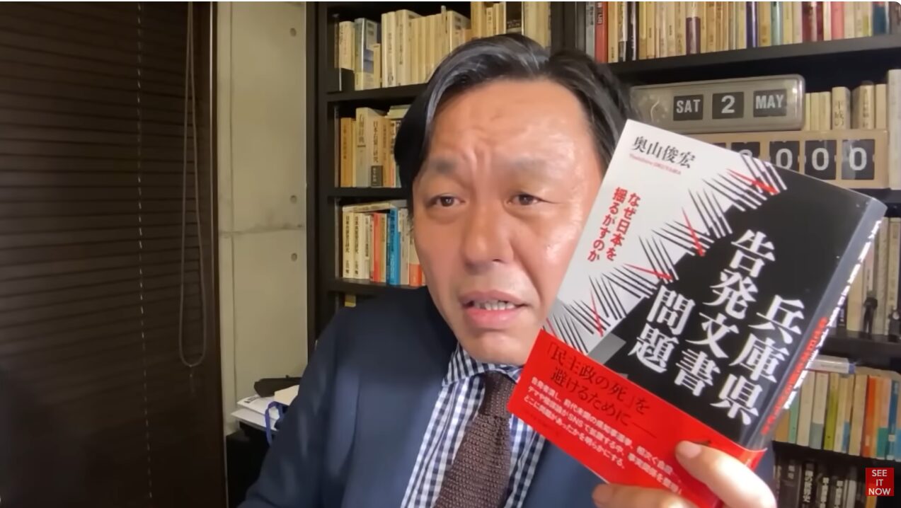 本棚を背に、奥山俊宏著『兵庫県告発問題書』を手に持ち、解説している菅野完。
