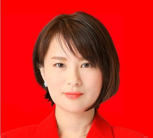鮮やかな赤色の背景を背に、ビジネススーツを着て凛とした表情で正面を見つめるショートヘアの深田萌絵。