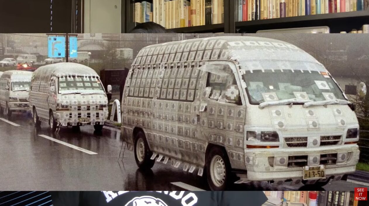車体全体に千円札のような模様の紙が無数に貼り付けられた、二台の白いワンボックスカーが雨の中を並走している様子。