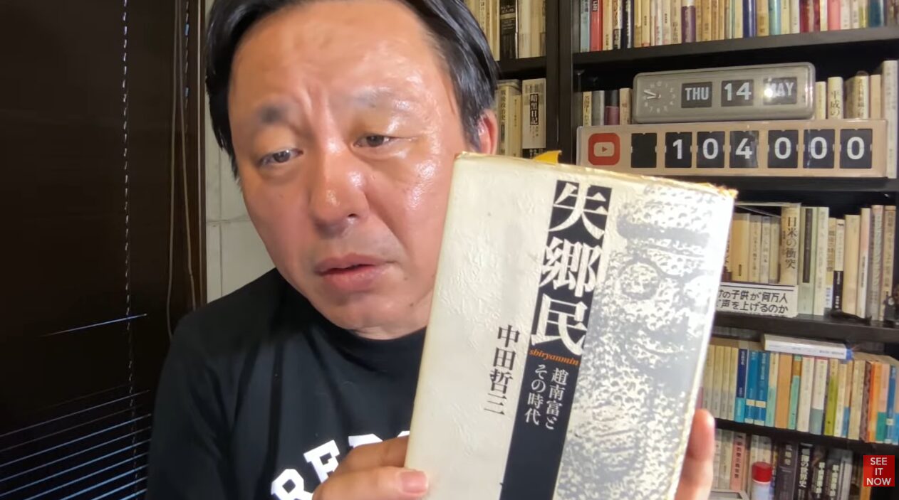本棚を背景に、中田哲三著『失郷民 趙南富とその時代』を手に持ち、内容を紹介する菅野完。