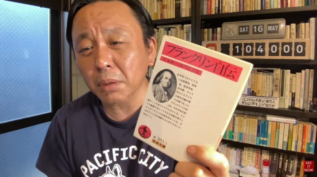 本棚の前で、手に持った岩波文庫『フランクリン自伝』をカメラに向けて紹介している菅野完。