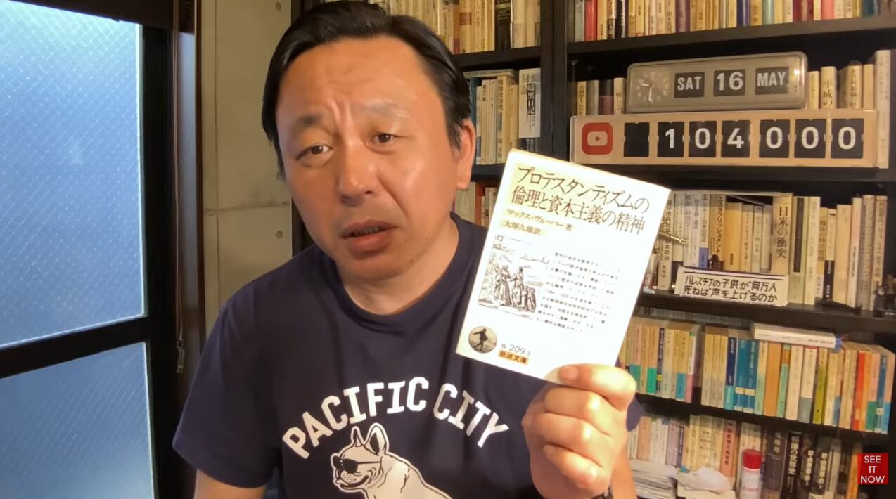 書籍『プロテスタンティズムの倫理と資本主義の精神』を手に持ち、解説している菅野完。背後には本棚がある。
