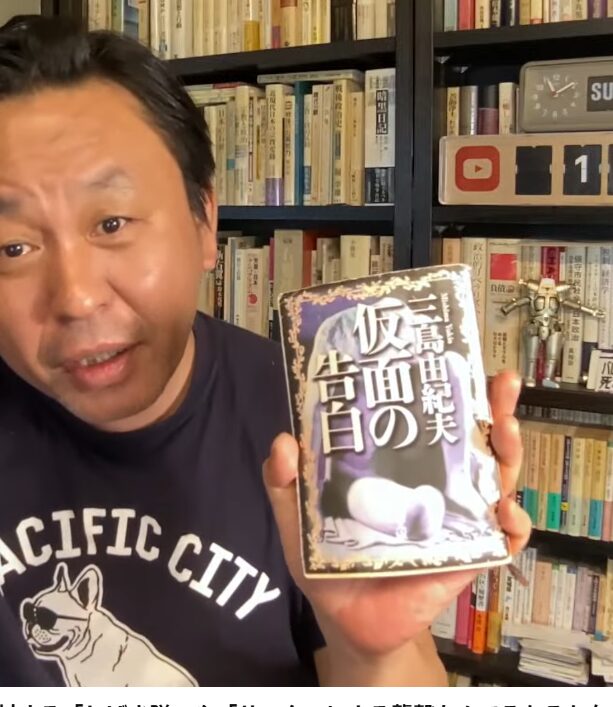 本棚を背にした菅野完が、三島由紀夫の小説『仮面の告白』を手に持ち、画面に向かって紹介している様子。