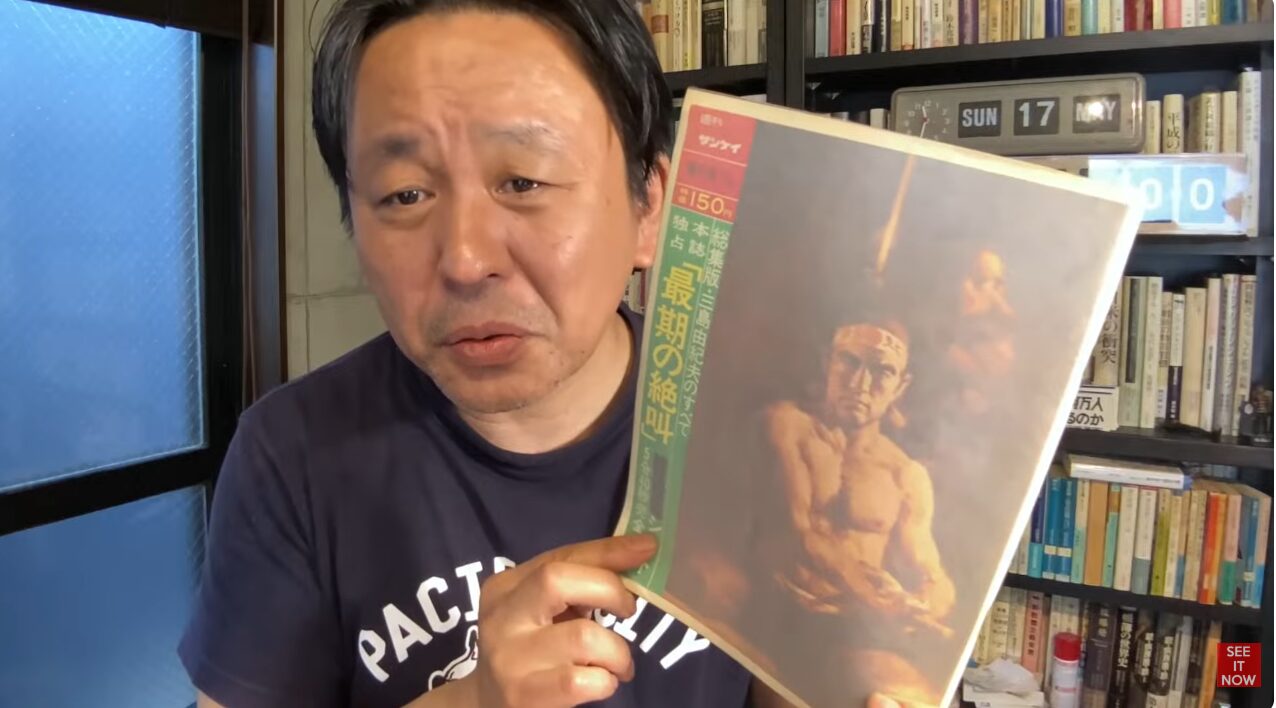 本棚を背にした菅野完が、三島由紀夫の特集が組まれた雑誌週刊サンケイを手に持って紹介している様子。