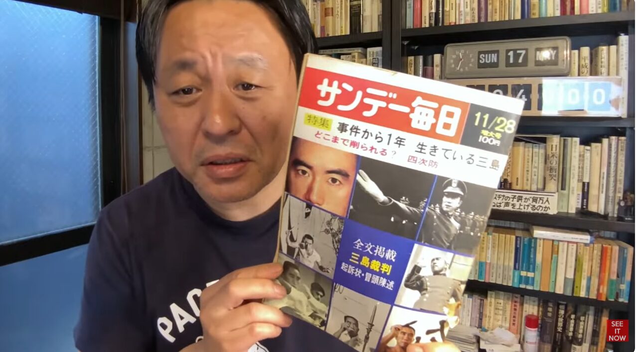 三島由紀夫特集が掲載された雑誌サンデー毎日を手に持ち、自身の顔の横で提示している菅野完。
