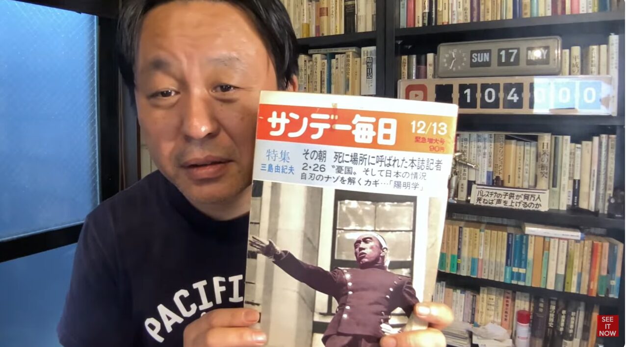 三島由紀夫を特集したサンデー毎日12月13日緊急増大号を手に持ち、解説する菅野完。背景には書籍が並ぶ棚がある。