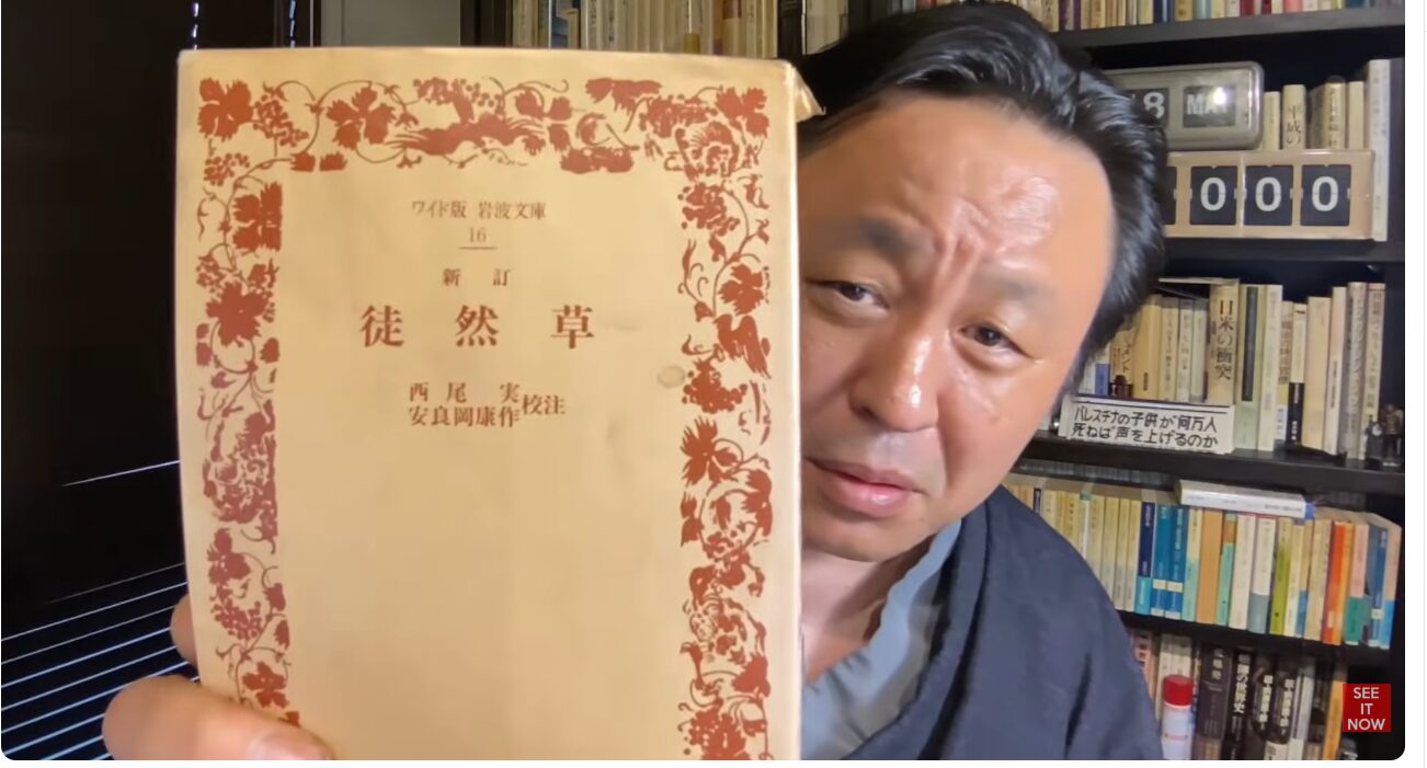 書棚を背にした男性が、ワイド版岩波文庫『新訂 徒然草』の表紙をカメラに向けて示している様子。