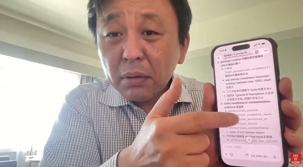 菅野完がスマートフォンの画面を指し示しながら、表示されている技術的な仕様やシステム設計に関するメモを解説している。