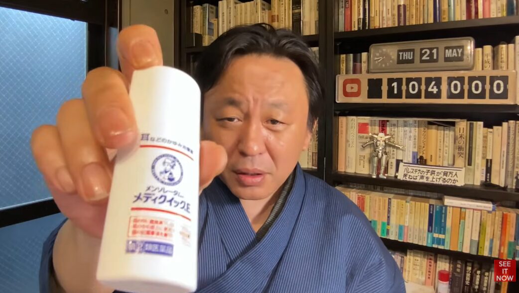 着物を着た菅野完が、本棚を背景にメンソレータム メディクイックEという耳のかゆみ止め薬をカメラに向けて提示している。