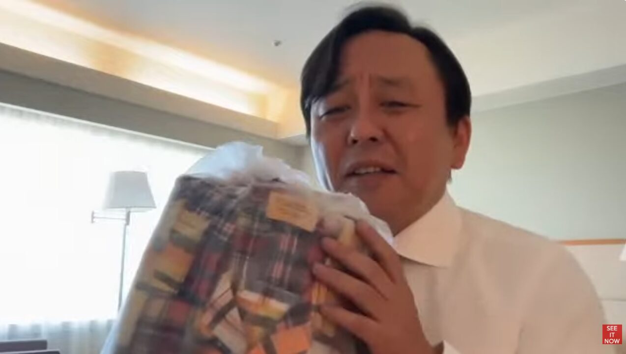 白いシャツを着た菅野完が、ホテルの部屋のような場所でチェック柄の衣類が入ったビニール袋を手に持ち、説明している様子。