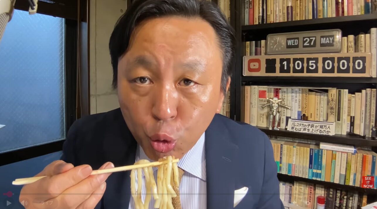 本棚を背景にスーツ姿の菅野完が箸で麺を持ち上げ、カメラに向かって食事をしている様子。