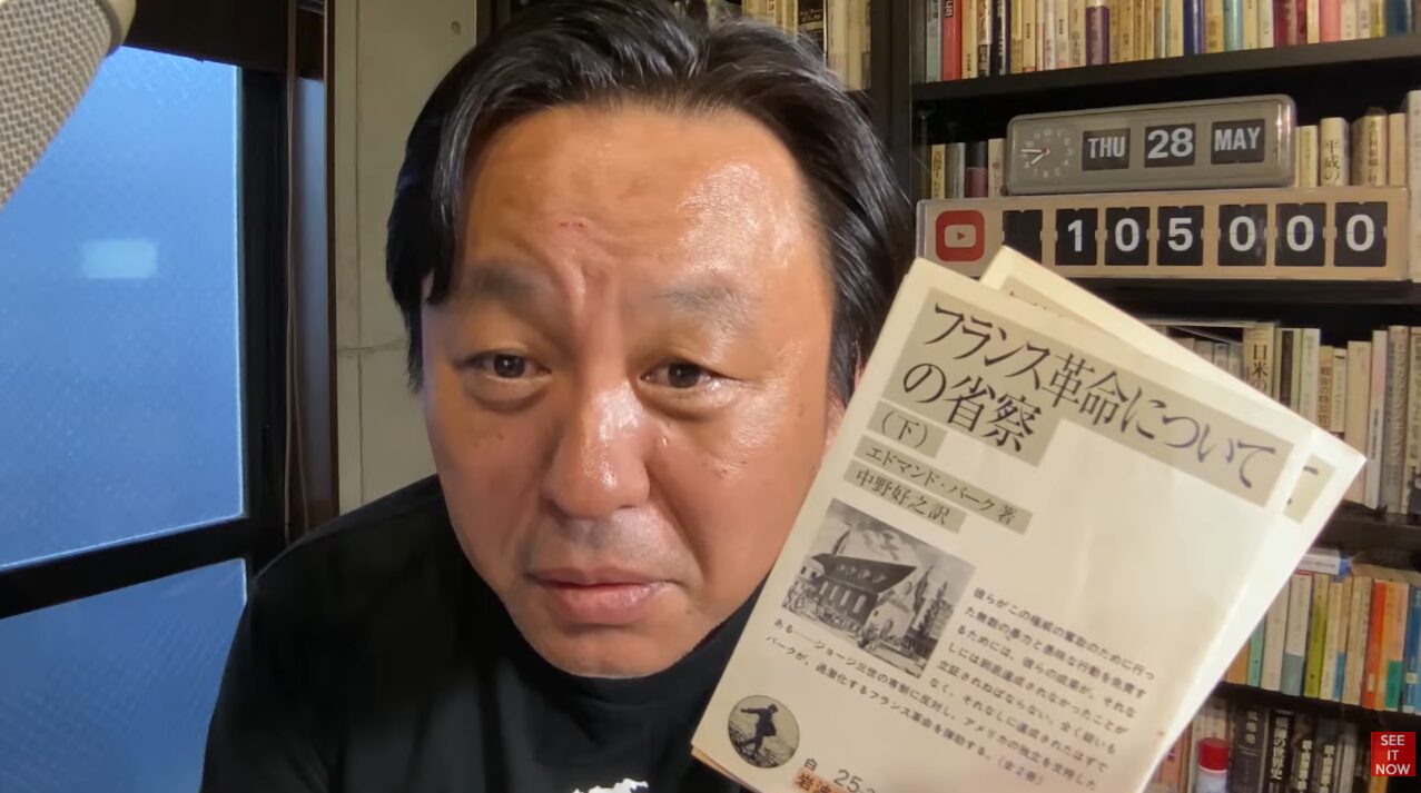 本棚を背にして『フランス革命の省察(下)』という書籍を手に持ち、カメラのレンズを見つめる菅野完。