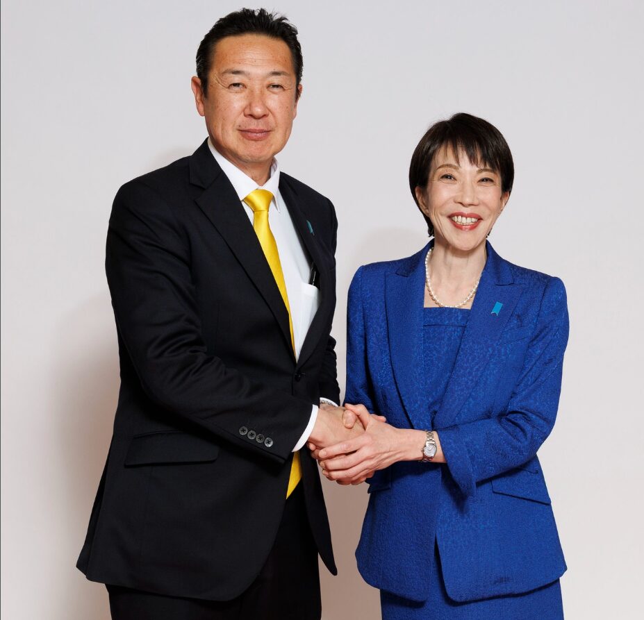 黄色のネクタイを締めたスーツ姿の黄川田仁志と、青いスーツを着用した笑顔の女性が並んで握手を交わしている。