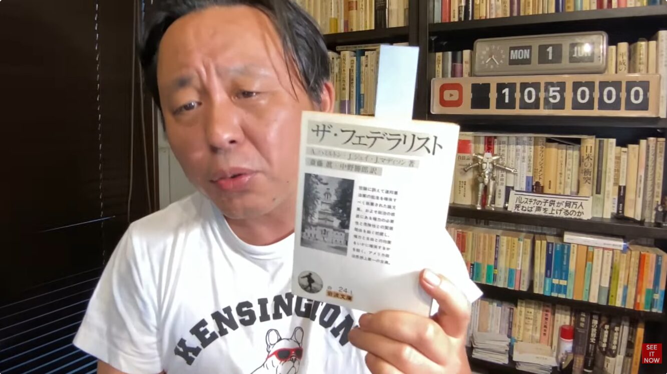 本棚を背にした菅野完が、手元で『ザ・フェデラリスト』という書籍を掲げて紹介している様子。