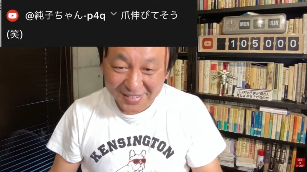 KENSINGTONのTシャツを着た菅野完が、本棚を背景にYouTubeライブ配信を行い、視聴者からの爪伸びてそう(笑)というコメントに反応している様子。