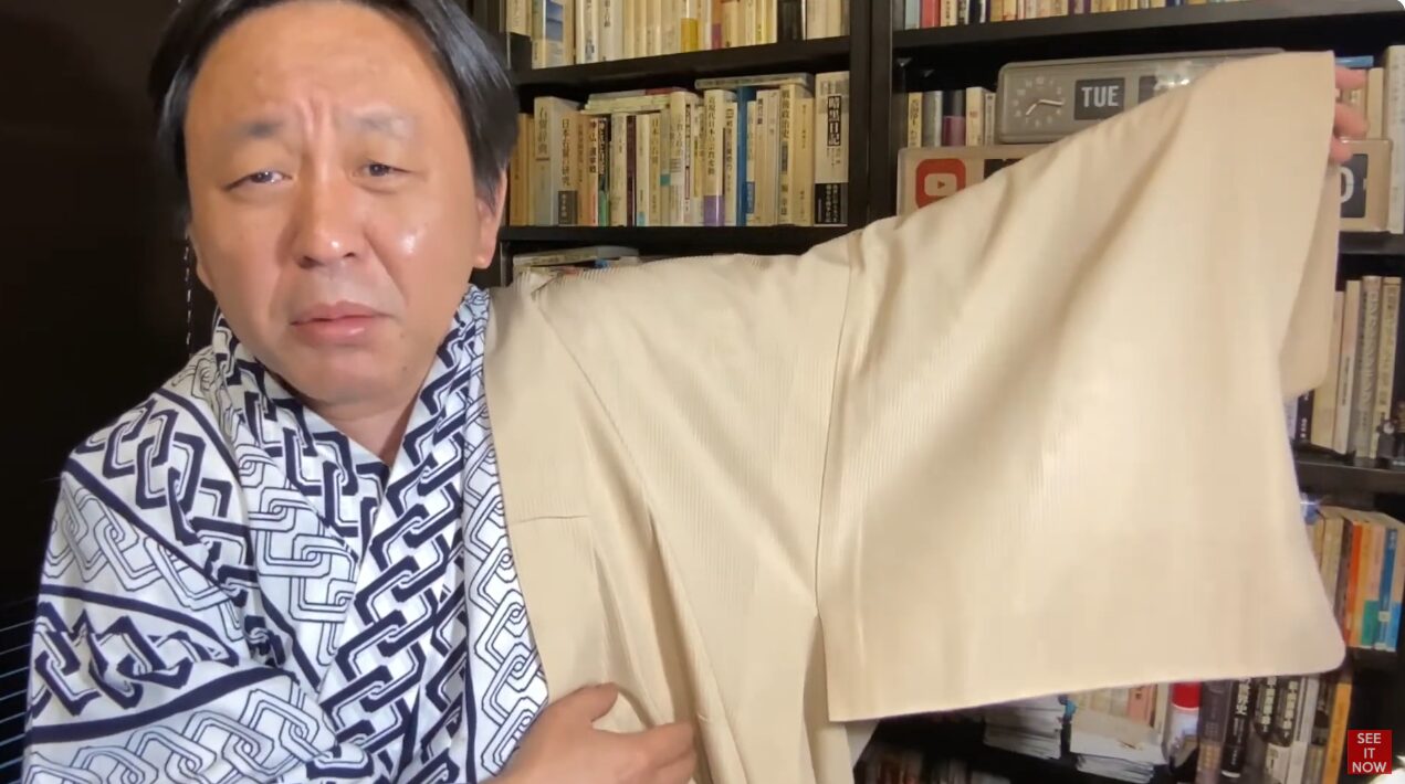 幾何学模様の着物を着た菅野完が、薄いベージュ色の着物の袖を広げて見せている様子。背景には書籍が並ぶ本棚がある。