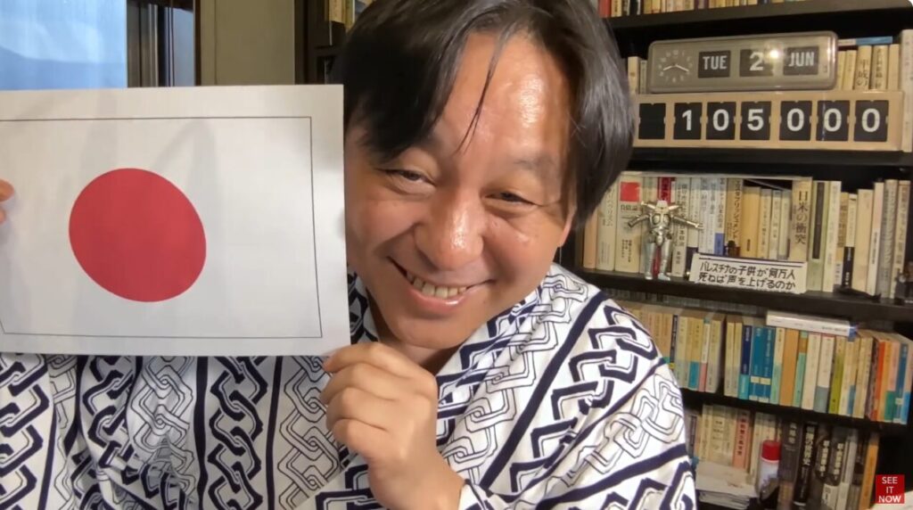 浴衣を着て笑顔で日の丸の紙を掲げる菅野完。背景には本棚があり、パレスチナの子供が何万人死ねば声を上げるのかと書かれた札が置かれている。