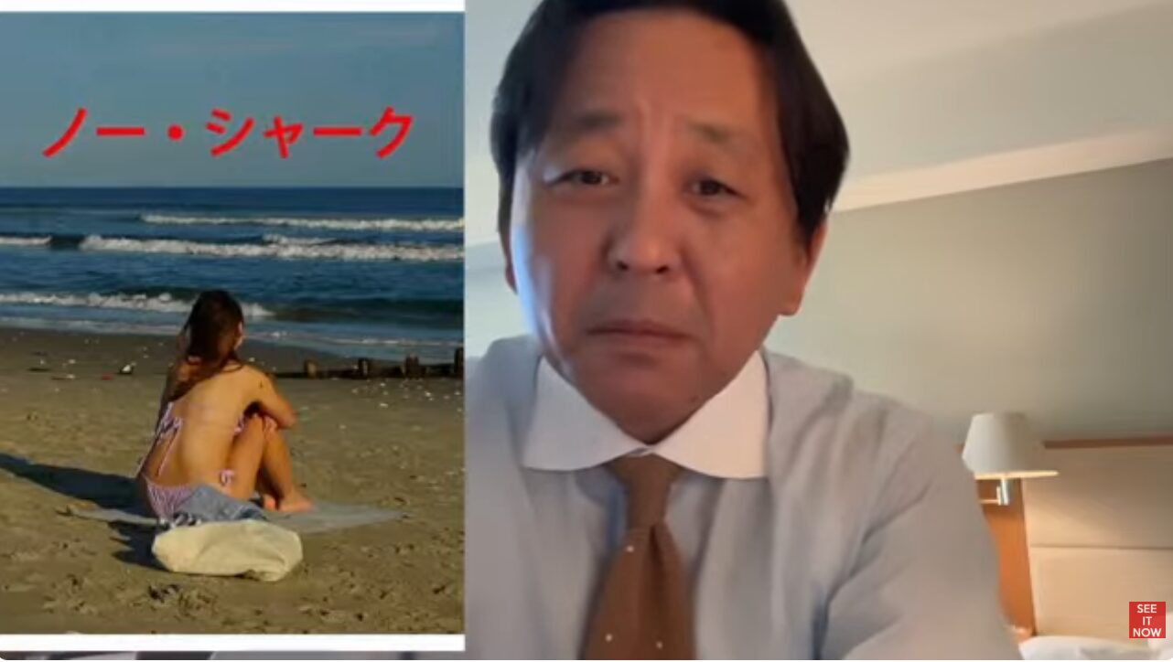 ノー・シャークの文字が重なる海辺で座る女性の映像を背景に、ネクタイ姿の菅野完が解説している様子。