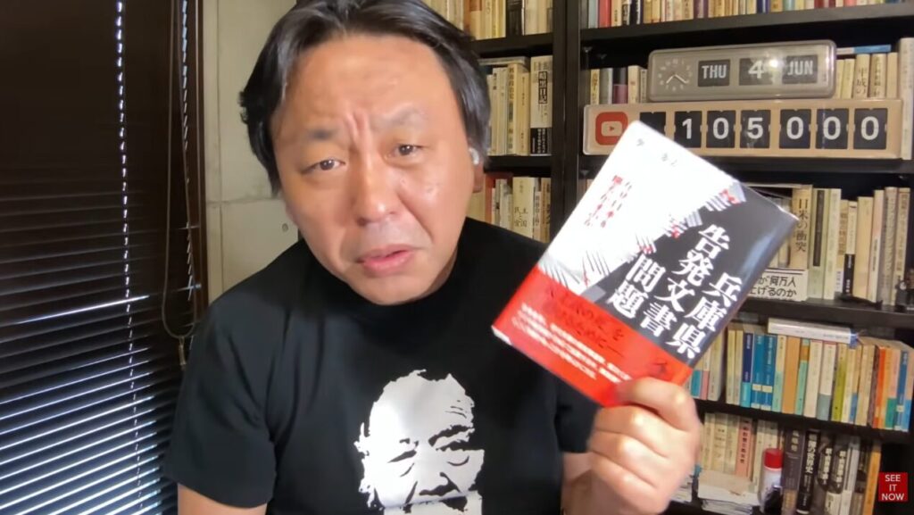 黒いTシャツを着た菅野完が、本棚を背景に『兵庫県告発文書問題』と記された書籍を手に持ち解説している。