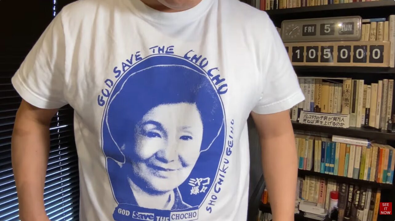 ミヤコ蝶々の肖像とGOD SAVE THE CHOCHOというロゴが青色でプリントされた白いTシャツを着た菅野完。背景には本棚が見える。