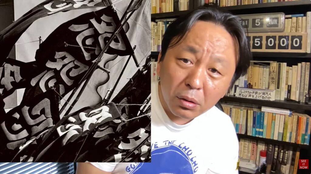 書棚の前で話す菅野完。画面左側には死と大きく書かれた旗が掲げられ、背景の棚にはパレスチナに関する書籍が置かれている。