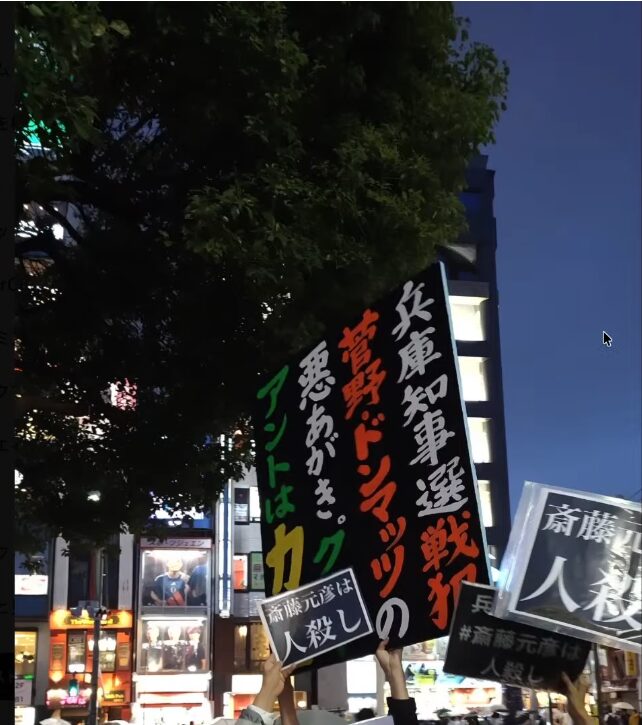 夜の街頭で、兵庫県知事選や斎藤元彦氏に対する批判的なメッセージが書かれたプラカードを掲げる様子。