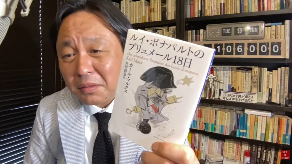 書棚の前で、カール・マルクスの著書『ルイ・ボナパルトのブリュメール18日』を手に持ち、解説するスーツ姿の菅野完。