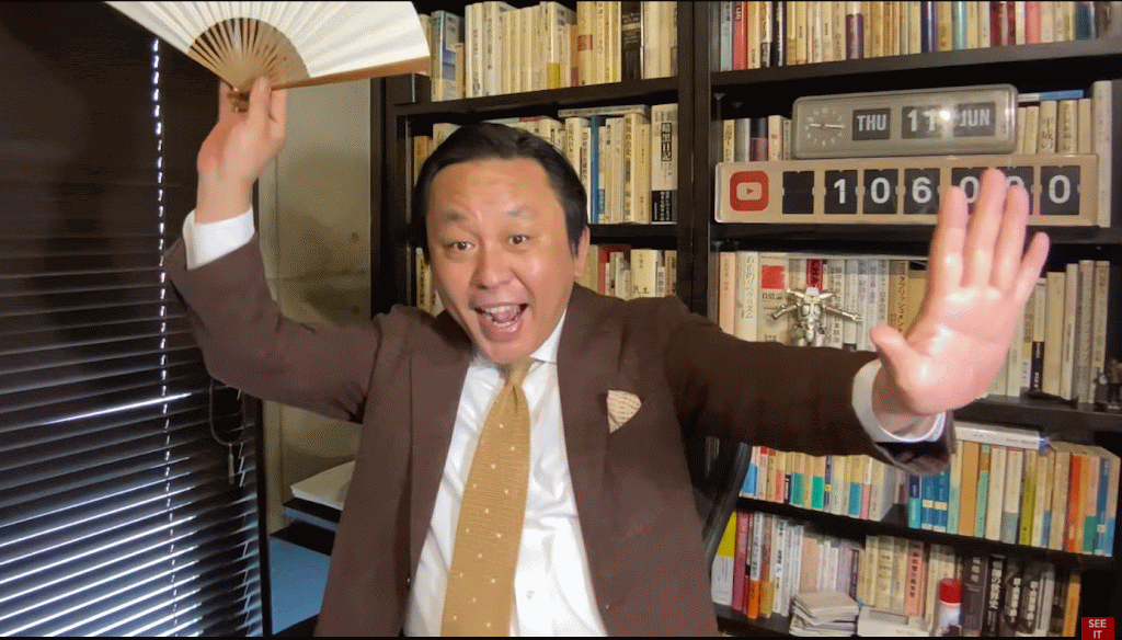 本棚を背景に、右手に扇子を持ち、左手を前に出して笑顔でポーズをとる茶色いジャケットを着た菅野完。