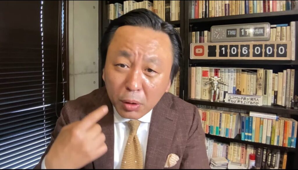 本棚を背景に、茶色のスーツを着た菅野完が口元に人差し指を向けながら、カメラに向かって熱心に語りかける。