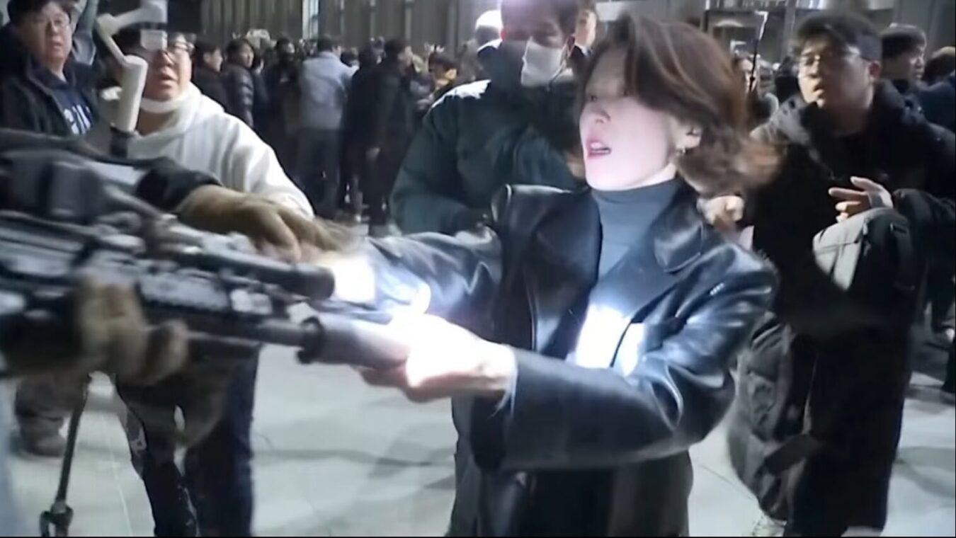 黒いレザージャケットを着た女性が、周囲の群衆の中でカメラマンの機材を押し返して道を切り開こうとしている様子。