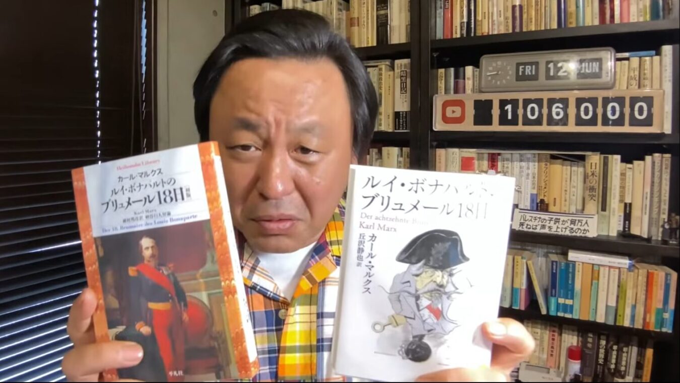 書棚の前で、カール・マルクス著『ルイ・ボナパルトのブリュメール18日』の異なる2つの版を手に持って紹介する菅野完。