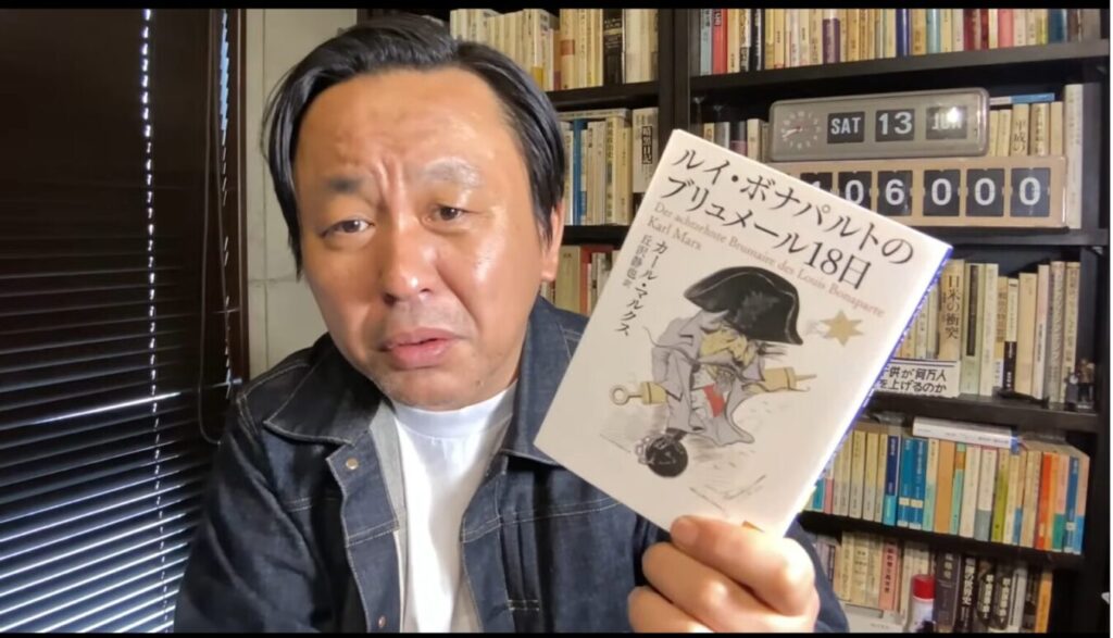 本棚を背景に、眉をひそめた菅野完がカール・マルクスの著書ルイ・ボナパルトのブリュメール18日を掲げる様子。
