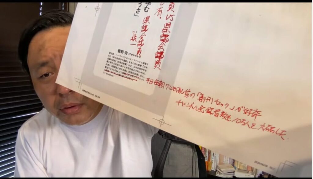 菅野完が自身のプロフィールや活動状況が記載された資料を掲げ、朝の配信番組の好調ぶりをアピールしている様子。