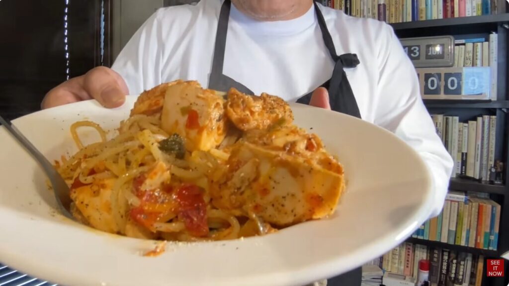 白いシャツとエプロン姿の菅野完が、トマトや具材をたっぷりと使用した手作りのパスタ料理を皿に盛り付けて差し出している様子。