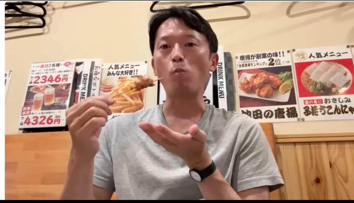 居酒屋の店内で、壁に貼られたメニューを背景に、斎藤元彦が焼き鳥の串焼きを口に運んで味わっている。