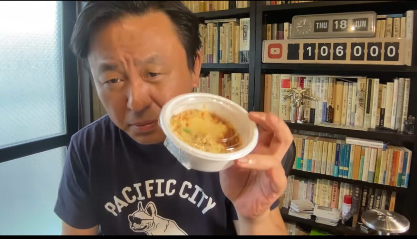 本棚を背景に、Tシャツ姿の菅野完が白いプラスチック容器に入った食べ物をカメラに向けて見せている。